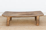 A rustic low wooden floor bench with a curved, thick seat and sturdy block legs, shown on a woven jute rug against a plain white wall.