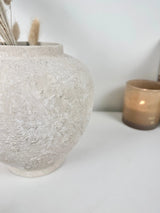 A textured, round ceramic vase in a light gray-beige finish, holding dried grass stems, set on a white surface with a softly blurred amber candle in the background.