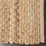 A close-up of a woven natural fiber rug or mat with vertical braided stripes in beige, tan, and light brown tones, shown on a dark wood surface. Callista Home