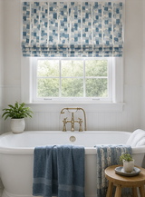A styled bathroom scene featuring a white classic bathtub with a brass faucet, draped with a blue striped towel, and a matching patterned blue and white Roman shade above a window. Callista Home