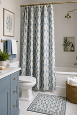 A bathroom scene showing a shower curtain with a blue and white geometric pattern, a matching bath mat, a blue vanity, a white toilet, and a wicker basket with towels. Callista Home