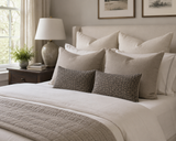 A neatly made bed in a softly lit bedroom, styled with multiple cream and beige pillows, two textured dark gray accent pillows, white bedding, a beige quilt, a dark wood nightstand, a table lamp, and framed wall art. Callista Home