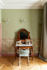 An ornate vintage wooden vanity table with a matching arched mirror, set against a sage green wall with floral wallpaper. A white chair with a light blue cushion sits in front of the vanity, which is styled with various small decorative items.