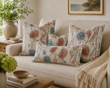 Three decorative throw pillows with a floral and geometric pattern in blue, coral, and beige, arranged on a cream-colored sofa in a cozy living room setting. Marcel 28A French botanical linen fabric by Antoinette Poisson Callista Home Callista Home