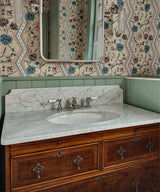 A vintage-style bathroom vanity featuring a white marble countertop with an integrated oval sink, antique bronze faucets, and a richly carved dark wood cabinet. The wall behind is decorated with ornate floral wallpaper and a sage green wainscoted panel, with a matching mirror reflecting the patterned wall.