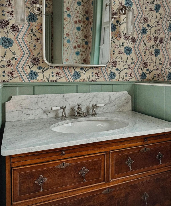 A vintage-style bathroom vanity featuring a white marble countertop with an integrated oval sink, antique bronze faucets, and a richly carved dark wood cabinet. The wall behind is decorated with ornate floral wallpaper and a sage green wainscoted panel, with a matching mirror reflecting the patterned wall.