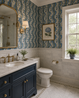 A styled bathroom interior featuring a navy blue vanity with gold hardware, a white marble countertop, a white toilet, and blue-and-white patterned wallpaper. A gold-framed floral print hangs on the wall, and a window on the right lets in natural light. Wallpapers Callista Home