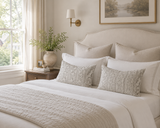 A neatly made bed in a softly lit bedroom, featuring white bedding, two textured beige accent pillows with a damask-style pattern, a quilted cream throw blanket, a wooden nightstand, a vase with greenery, and a wall sconce. Callista Home