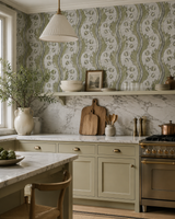 A styled kitchen interior featuring sage green cabinets, a white marble countertop, a vintage-style cream pendant light, and a wall covered in green and white wavy botanical wallpaper. Callista Home