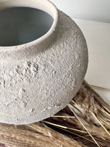 A textured, matte gray ceramic pot with a rough, hand-painted finish, resting on dried pampas grass against a clean white background.