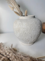 A textured concrete-style ceramic vase in a light gray color, styled with dried pampas grass on a white surface against a neutral gray background.