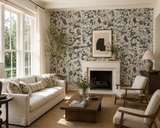 A styled living room featuring a prominent black and white abstract floral wallpaper on the wall behind a white fireplace mantel. The room is furnished with a cream-colored sofa, two matching armchairs, a dark wood coffee table, and a large window on the left.