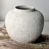 A round, textured ceramic vase in a light warm gray or off-white tone, styled on a surface with dried pampas grass against a neutral background.