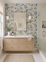 A modern bathroom vanity setup with a light wood floating cabinet, white countertop, gold faucet, and a large mirror set against a blue and beige floral wallpaper. A glass shower door is visible on the right, and a woven rug sits on the floor. Jaipur 57B blue French wallpaper by Antoinette Poisson featuring a historic domino paper pattern with an ultra-matte finish. Callista Home