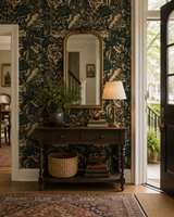 An ornate interior entryway featuring dark green and beige floral wallpaper, a dark wood console table with a mirror, a table lamp, and decorative baskets, with a doorway on the right leading outside. Grands Pavots 59A large-scale floral poppy French wallpaper Callista Home