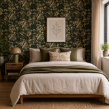A styled bedroom featuring a bed dressed in cream and olive bedding, set against a dark green floral wallpaper wall with a framed botanical print above. A wooden nightstand with a lamp sits on the left, and a window with sheer curtains is on the right. Callista Home