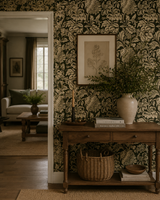 A styled interior scene featuring dark green and beige floral patterned wallpaper, a wooden console table with a cream ceramic vase of greenery, books, and a wicker basket, with a doorway leading to a softly lit living room. Callista Home