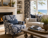 A beautifully styled coastal living room featuring a navy blue and cream floral patterned armchair next to a cream sofa, with a large window overlooking the blue ocean. Callista Home