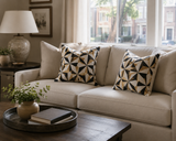 A beige upholstered sofa in a living room setting, styled with two square throw pillows featuring a black, white, and beige geometric starburst pattern. A dark wood coffee table with a tray and books sits in the foreground, and a lamp and window are visible in the background. Callista Home