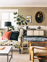 A styled living room interior featuring a floral sofa with red and black accent pillows, a tall potted fiddle-leaf fig plant, a patterned wallpapered wall with framed art, a white sideboard, and a wooden armchair in the foreground. Callista Home