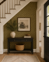 An entryway scene with a black console table against a wall covered in olive-green and black geometric patterned wallpaper. A white vase with greenery, books, a framed landscape print, and a woven basket sit on the table. Alvéoles Wallpaper 60B by Antoinette Poisson featuring a refined repeating geometric pattern inspired by historic 18th-century French domino paper. Callista Home