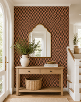 A styled interior entryway featuring a brown and beige patterned wallpapered accent wall, a light wood console table, a white ceramic vase with greenery, a wicker basket, and a gold-framed mirror. Alvéoles Wallpaper 60C by Antoinette Poisson featuring a repeating geometric motif inspired by historic 18th-century French domino paper. Callista Home
