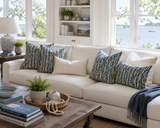 A white sectional sofa in a bright living room styled with four patterned throw pillows in blue, yellow, and white, along with cream pillows and a dark blue throw blanket. A wooden coffee table with books and a plant sits in the foreground, and a window in the background shows water and trees. Callista Home