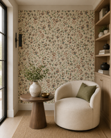 A styled interior corner featuring a cream upholstered accent chair beside a round wooden side table with a white ceramic vase and greenery, set on a woven rug against a wall covered in dense vintage floral wallpaper. A light wood shelving unit stands on the right, and a window frame is visible on the left. Callista Home