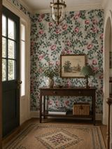 A classic entryway featuring floral wallpaper with pink roses and green leaves on a light blue-gray background. A dark wood console table sits against the wall, decorated with a vase of flowers, a green lamp, and a framed landscape print. Callista Home.