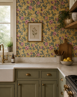 A kitchen interior with yellow floral wallpaper, sage green cabinets, a white farmhouse sink, brass fixtures, and a framed botanical print on the wall. Callista Home
