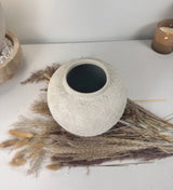 A round, textured off-white ceramic pot with a dark interior, styled on a white tabletop with dried pampas grass and a lit candle in the background.