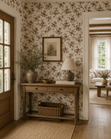 A styled interior corner with floral wallpaper, a wooden console table, a lamp, framed wall art, and a basket beneath the table. An open doorway on the right reveals a sunlit living room with a sofa and coffee table.
