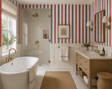 A bright, modern bathroom featuring bold red, white, and blue vertical striped wallpaper, a white freestanding bathtub with gold fixtures, a wood vanity, and a glass shower enclosure.