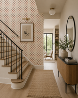 A styled interior hallway with a staircase on the left, patterned wallpaper, a woven rug, a wood console table with a black countertop, and a large oval mirror on the right.