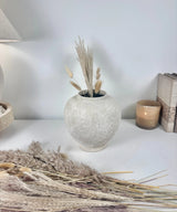 A textured, round ceramic vase in a light beige-gray color, styled with dried pampas grass on a white tabletop against a light gray wall.