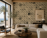 A styled living room interior featuring a large wall covered in repeating vintage-style illustrated tiles in beige and black. The room includes a cream sectional sofa, a round wood coffee table, a black accent chair, a palm tree in a large white planter, and a large framed art print on the wall.