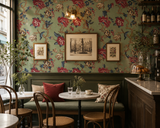 A cozy cafe interior with green floral wallpaper, a green upholstered bench with red and patterned pillows, small round tables, wooden chairs, framed wall art, and a marble counter.