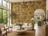 A styled living room featuring a bold mustard yellow wallpaper with a dense red, pink, and blue floral pattern on the main wall. The room is furnished with a cream sofa, a round stone coffee table, wooden chairs, and large windows looking out onto green trees.