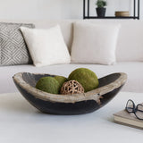 A rustic dark bowl with a natural wood rim, styled on a white table with moss balls and a woven ball inside, set in a bright living room with a sofa and pillows in the background. Callista Home