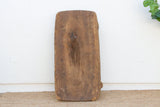 A rustic, rectangular wooden board with rounded corners and a prominent natural wood grain pattern, standing upright on a woven jute rug against a white wall.