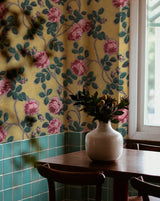 Floral wallpaper with pink roses and green leaves on a yellow background, paired with a white vase on a wooden table. Callista Home