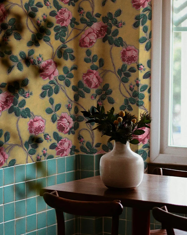 Floral wallpaper with pink roses and green leaves on a yellow background, paired with a white vase on a wooden table. Callista Home