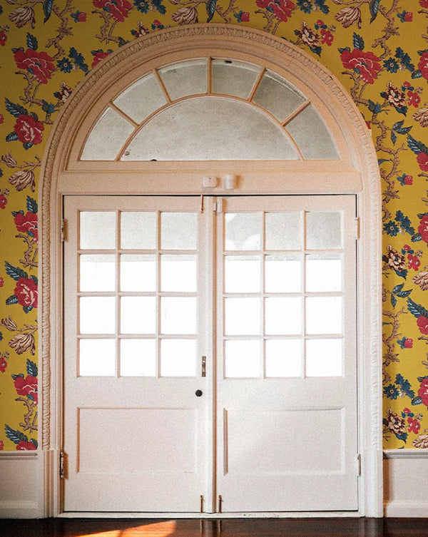 A pair of white double doors with multiple square glass panes and a decorative arched transom window above, set against a bright yellow floral wallpaper.