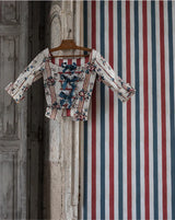 A long-sleeve blouse with a red, white, and blue striped pattern and blue bow details on the front, hanging on a wooden hanger against a rustic wooden door and a red, white, and blue striped wall.