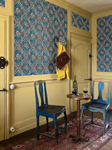 Vintage room with blue chairs, a small table, and patterned wallpaper. Guirlandes de Fleurs 1A blue floral French wallpaper CAllista Home