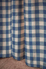 Blue and off-white gingham checkered curtain panels hanging over a window, with a textured brown carpet visible at the bottom. Carreaux Indigo 84A blue geometric linen cotton fabric by Antoinette Poisson Callista Home