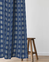 A blue patterned curtain panel hanging next to a wooden stool in a minimalist room setting with a white wall and light wood floor. Flammé Indigo 83A indigo blue geometric French linen cotton fabric Callista Home