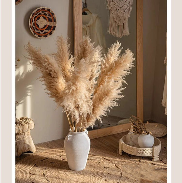 Decorative pampas grass in a white vase on a wooden surface with a mirror and wall art in the background. Callista Home