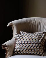 A square throw pillow with a geometric triangle pattern in beige, black, and white, resting on a distressed cream-colored armchair against a dark background. Callista Home