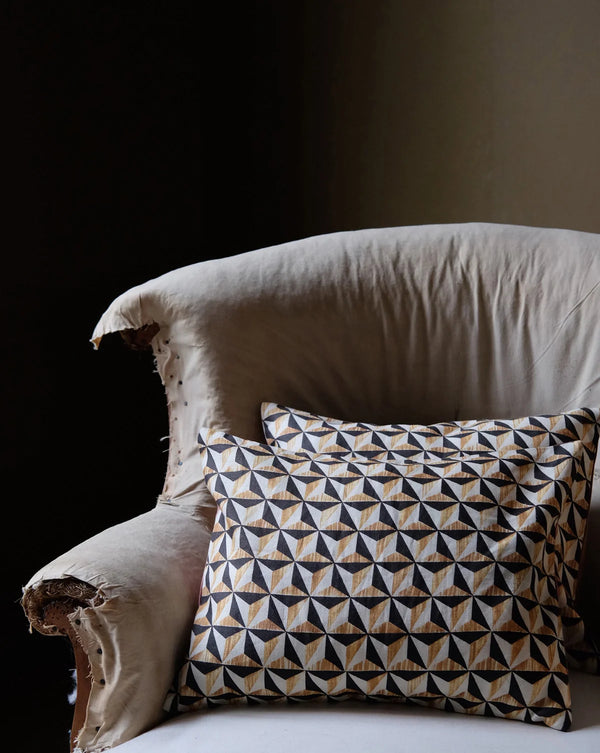 A square throw pillow with a geometric triangle pattern in beige, black, and white, resting on a distressed cream-colored armchair against a dark background. Callista Home